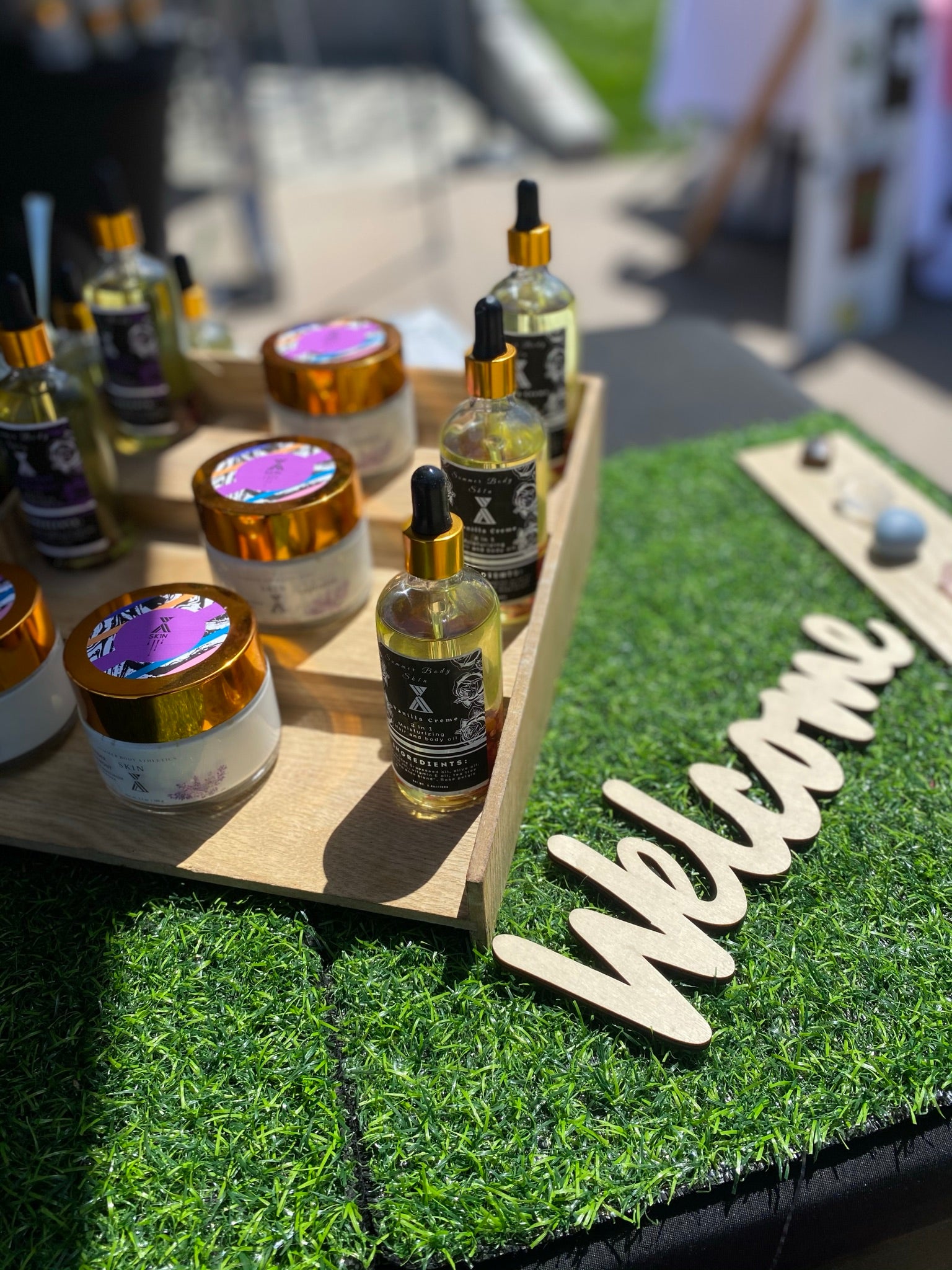 A vendor table covered with a grass top and three tier stairs to display cosmetics. Each bertical row displays a beautiful gold glass bottle containing scent body care made with all natural ingredients. The packaging design is colorful, clean, and culturally authentic.